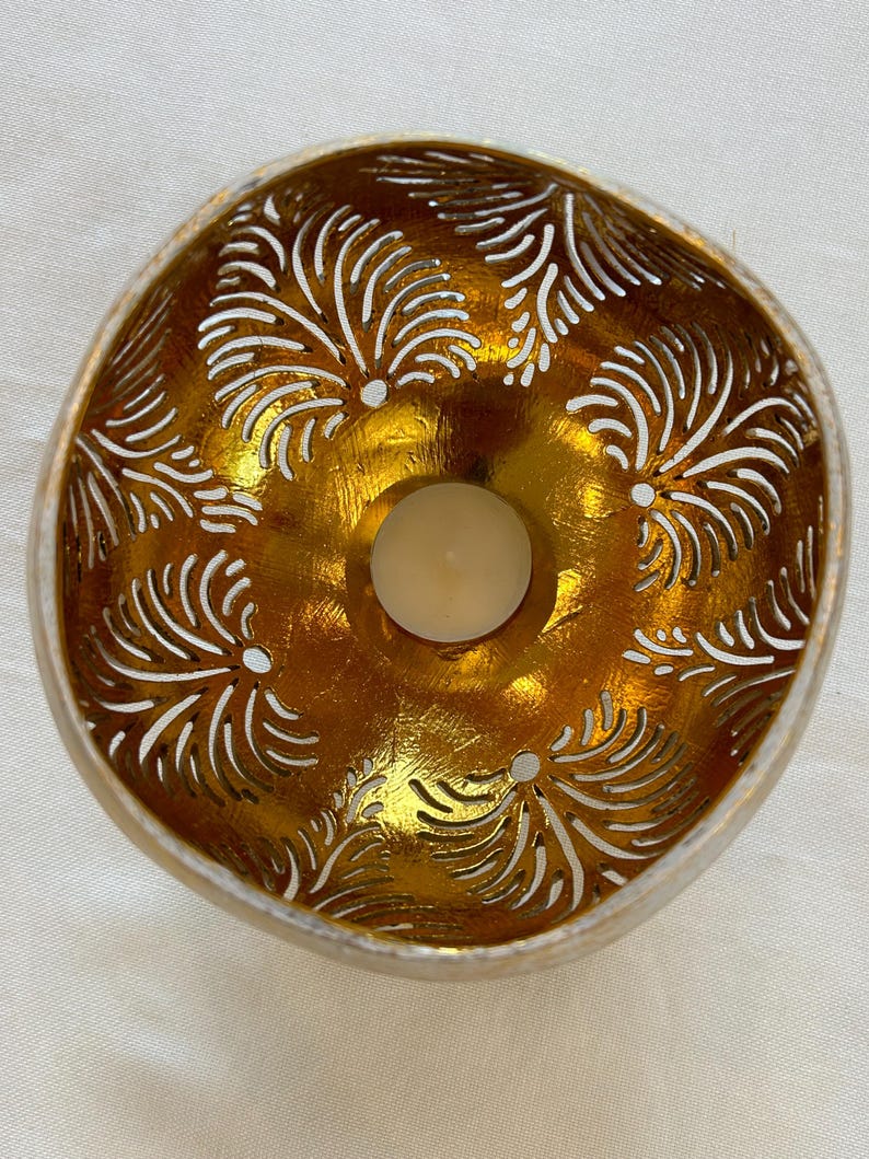 May include: A gold-painted coconut shell candle holder with a white, intricate, carved design. The holder has a small, round opening in the center for a tea light candle.