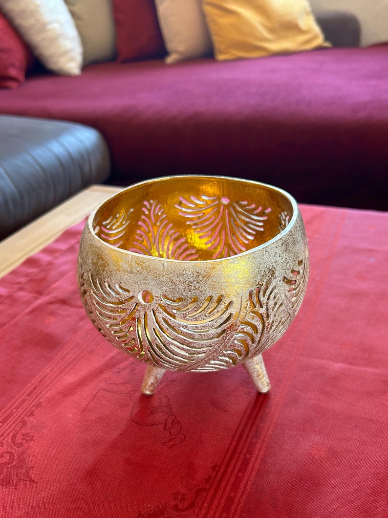 May include: Gold metal candle holder with a decorative cut-out design. The holder has three legs and is sitting on a red table cloth.