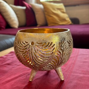 May include: A gold metal candle holder with a decorative leaf pattern and three legs. The candle holder is sitting on a red tablecloth.