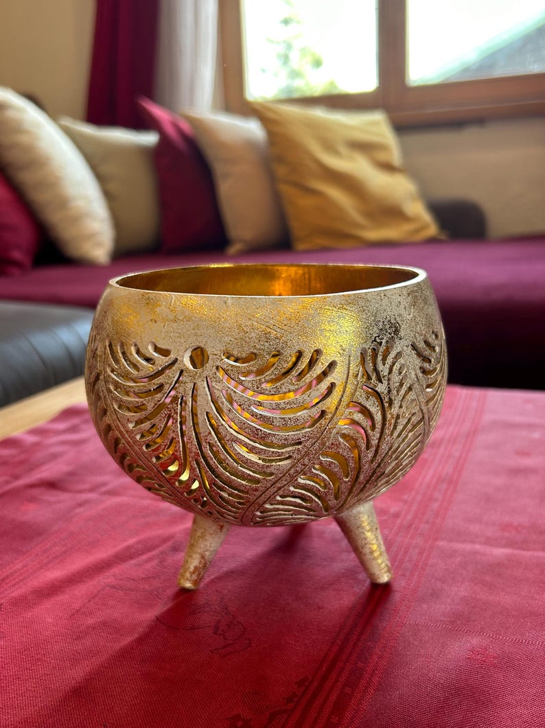 May include: A gold metal candle holder with a decorative leaf pattern and three legs. The candle holder is sitting on a red tablecloth.