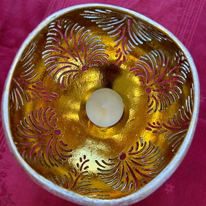 May include: A white coconut shell bowl with a gold leaf design and a small white candle in the center. The bowl is decorated with a carved pattern of leaves.