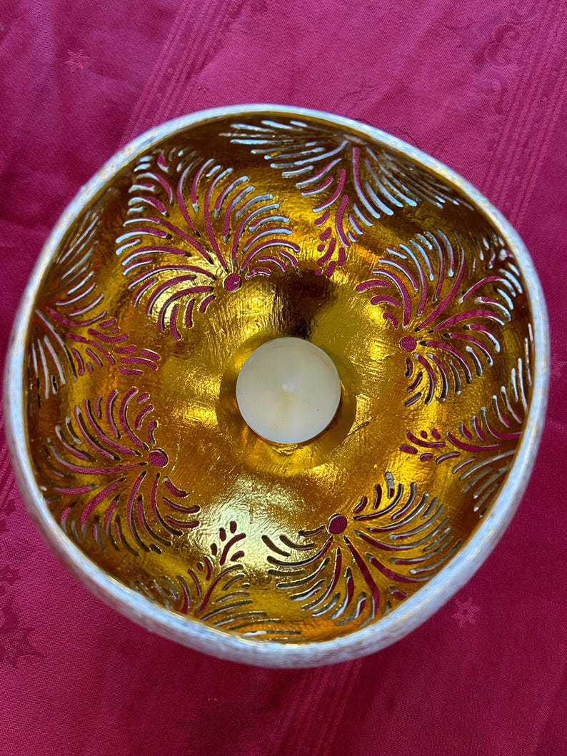 May include: A white coconut shell bowl with a gold leaf design and a small white candle in the center. The bowl is decorated with a carved pattern of leaves.