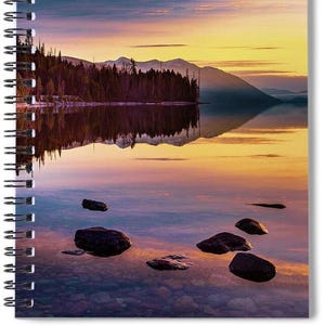 May include: Spiral-bound notebook featuring a scenic landscape. The image showcases a lake reflecting a colorful sunset with hues of orange, yellow, and purple. Dark rocks are scattered in the foreground, and a forest lines the water's edge.