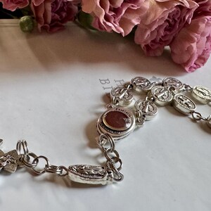 May include: A silver rosary with detailed religious medallions and a cross. The rosary features a central bead with the word "Jerusalem" inscribed. Pink flowers are in the background.