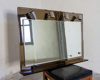 May include: A large, rectangular mirror with a brown frame and four chrome light fixtures. The mirror is mounted on a wall and has a shelf underneath.