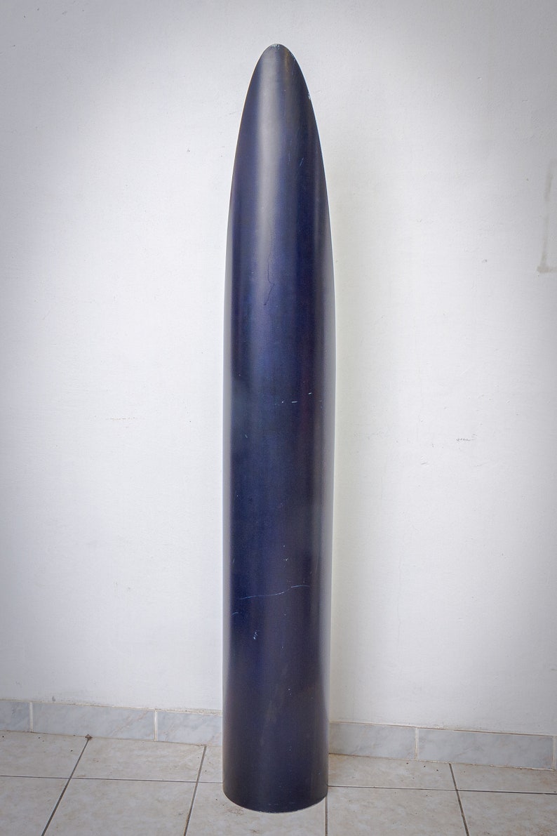 May include: A tall, dark blue, cylindrical object with a pointed top. The object is standing upright against a white wall and a tiled floor. The surface of the object appears smooth with some minor imperfections.