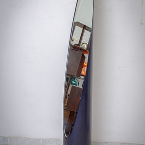May include: A tall, navy blue, oval-shaped floor mirror. The mirror is encased in a cylindrical frame. The mirror reflects a room with wooden shelves and various objects. The mirror is a decorative home decor item.