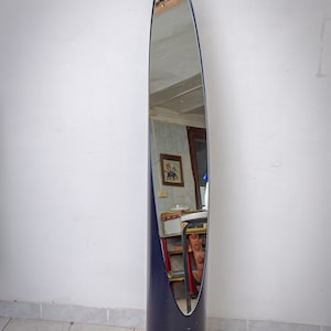 May include: A tall, oval-shaped floor mirror with a dark blue frame and base. The mirror's reflective surface is surrounded by a thin blue border. The base is cylindrical, and the mirror's shape is reminiscent of a surfboard.