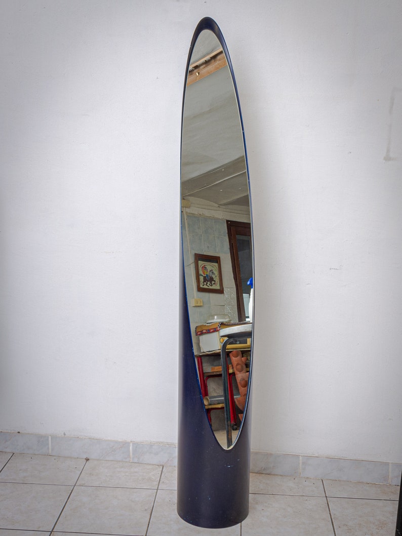 May include: A tall, oval-shaped floor mirror with a dark blue frame and base. The mirror's reflective surface is surrounded by a thin blue border. The base is cylindrical, and the mirror's shape is reminiscent of a surfboard.