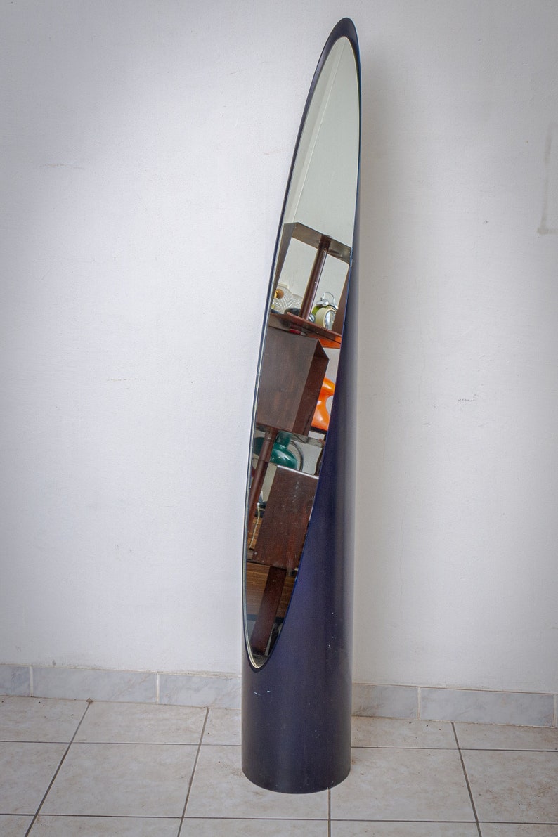 May include: A tall, navy blue, oval-shaped floor mirror. The mirror is encased in a dark blue frame and has a sleek, modern design. The mirror reflects the surrounding room, including furniture and decor.