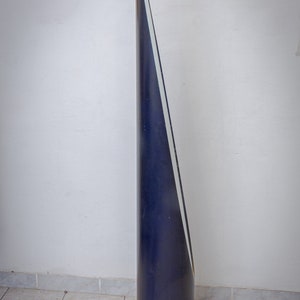 May include: A tall, dark blue, conical sculpture with a white stripe running along one side. The sculpture is cylindrical at the base and tapers to a sharp point. It stands on a tiled floor against a white wall.