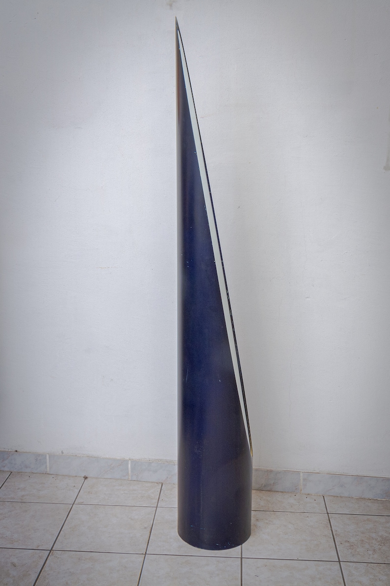 May include: A tall, dark blue, conical sculpture with a white stripe running along one side. The sculpture is cylindrical at the base and tapers to a sharp point. It stands on a tiled floor against a white wall.