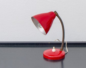 May include: A vintage red desk lamp with a flexible, gooseneck arm and a round base. The lamp has a conical shade with a silver interior and a white on/off switch. The lamp is on a black surface.