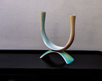May include: A ceramic candle holder with two arms in the shape of a U. The holder is a light blue and yellow color with a white base.