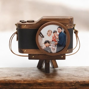 May include: A wooden camera-shaped photo frame featuring a dark brown wood body with black textured panels, gold-tone metal accents, and a circular lens-style frame displaying a photograph of a family. The frame rests on a small wooden tripod stand.