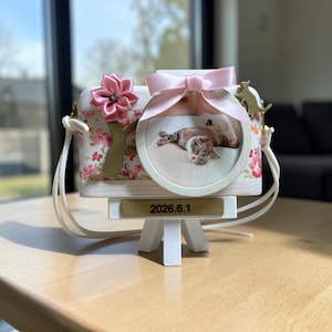 以下が含まれることがあります： 花柄の布パネル、ピンクのリボンの結び目、布製の花、猫の丸い写真が特徴の白い木製のカメラ型フレーム。金色のプレートには「2026.6.1」という文字が表示されています。フレームは小さな白い木製のイーゼルスタンドの上に置かれています。