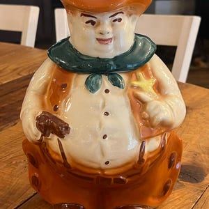 May include: A ceramic cookie jar shaped like a cowboy figure wearing a brown hat, a dark green neckerchief, and orange trousers. The figure has a cream-colored shirt with brown buttons, a yellow star-shaped badge, and holds a small brown object.