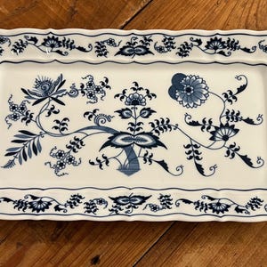 May include: A rectangular white ceramic serving tray with scalloped edges and integrated handles. The surface features a detailed blue floral pattern with stylized blossoms, leaves, and vines arranged in a symmetrical composition.