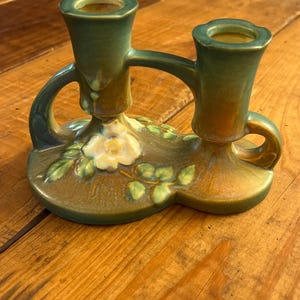 May include: A dual-holder ceramic candle holder featuring a glazed finish in shades of forest green and amber. The base is decorated with a sculpted white flower and green leaves, with two cylindrical sockets connected by a curved bridge handle.