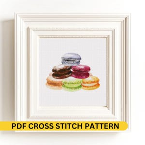 Op de afbeelding: Een ingelijst kruissteekpatroon met een stapel kleurrijke macarons. De macarons zijn in tinten bruin, roze, groen, oranje en blauw. De lijst is wit en de tekst "PDF CROSS STITCH PATTERN" staat onderaan.