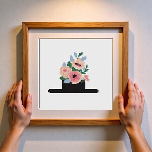 Op de afbeelding: Kruissteekkunst van een zwarte hoed die overloopt van roze en perzik bloemen en blauwe bladeren. Het kunstwerk is ingelijst in een lichtbruine houten lijst, vastgehouden door twee handen.