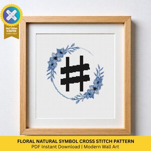 May include: A framed cross-stitch design with a black hashtag symbol surrounded by a wreath of blue flowers and foliage. The artwork is set against a white backdrop and framed in light wood. Text at the bottom reads "FLORAL NATURAL SYMBOL CROSS STITCH PATTERN".