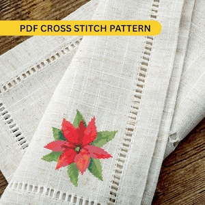 May include: A folded, off-white linen fabric with a cross-stitched poinsettia design. The red and green floral pattern is centered on the fabric. The text "PDF CROSS STITCH PATTERN" is in a yellow banner at the top.
