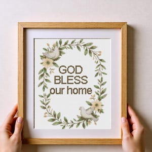 Puede incluir: Una obra de arte en punto de cruz enmarcada con las palabras "GOD BLESS our home" en letras marrones. El texto está rodeado por una corona de hojas verdes, flores amarillas y dos palomas blancas. El marco es de madera marrón claro.