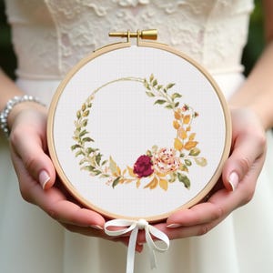 May include: A cross-stitch embroidery hoop featuring a floral wreath design. The wreath includes green leaves, yellow and orange foliage, and pink and burgundy flowers. The embroidery is set in a wooden hoop with a gold clasp and a white ribbon.