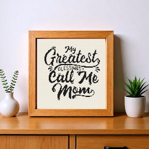 May include: A framed cross-stitch artwork with the text "My Greatest Blessings Call Me Mom" in black script. The artwork is in a light-coloured frame, set on a wooden surface, with small plants in white vases on either side.