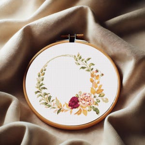 Puede incluir: Un aro de bordado con fondo blanco, que presenta un diseño de corona de flores con flores de color burdeos y melocotón y hojas amarillas.