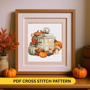 May include: Cross-stitch pattern framed in a light wood frame. The artwork features a white lantern surrounded by pumpkins, fall leaves, and a mushroom. The text "PDF CROSS STITCH PATTERN" is displayed on a yellow banner at the bottom.