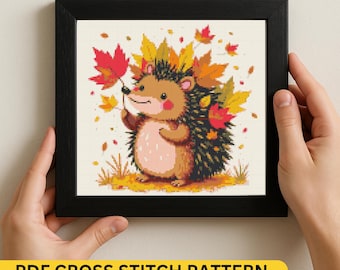 May include: A framed cross-stitch pattern featuring a cartoon hedgehog surrounded by autumn leaves. The hedgehog is brown, tan, and pink, holding a red leaf. The frame is black, and the text "PDF CROSS STITCH PATTERN" is at the bottom.