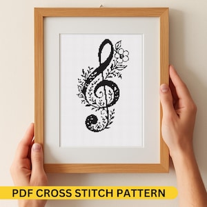 Può includere: Un modello a punto croce incorniciato con un disegno di chiave di violino nera adornato da elementi floreali. L'opera d'arte è presentata in una cornice di legno con un passe-partout bianco. Il testo "PDF CROSS STITCH PATTERN" è visibile in basso.