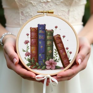 May include: A cross-stitch embroidery hoop featuring a stack of colorful books with floral accents. The books are in shades of red, blue, and green, with pink flowers and green leaves. The hoop is held by hands.