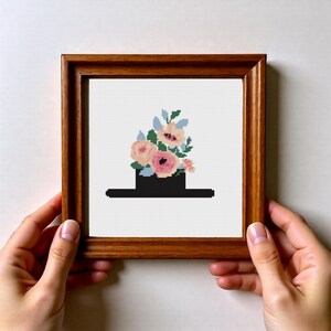 Op de afbeelding: Een ingelijst kruissteekkunstwerk met een zwarte hoed met een boeket van roze en perzik bloemen, geaccentueerd met blauwe bladeren. Het kunstwerk is in een bruine houten lijst.