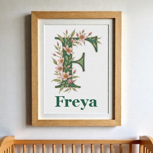 Op de afbeelding: Een ingelijst kruissteek kunstwerk met een grote, donkergroene letter F versierd met roze bloemen en groene bladeren. Onder de letter is de naam Freya geborduurd in donkergroene hoofdletters. Het werk zit in een lijst van lichtgekleurd hout.