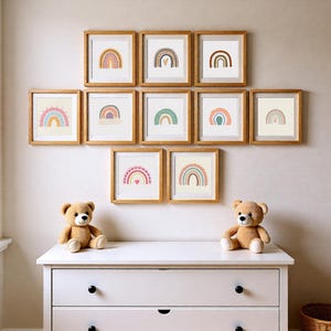 May include: A display of framed rainbow art prints in a variety of colours, mounted on a wall. The prints are in wooden frames. Two teddy bears sit on a white chest of drawers below the artwork.