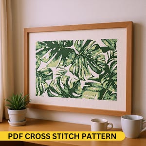 Könnte beinhalten: Ein gerahmtes Kreuzstichmuster mit einem Design aus grünen Monstera-Blättern. Das Kunstwerk befindet sich in einem hellbraunen Holzrahmen. Der Text "PDF CROSS STITCH PATTERN" befindet sich unten.