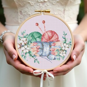 May include: A cross-stitch embroidery hoop featuring a design of yarn balls and knitting needles in shades of green, pink, and blue, surrounded by floral accents. The wooden hoop is held with a white ribbon bow.