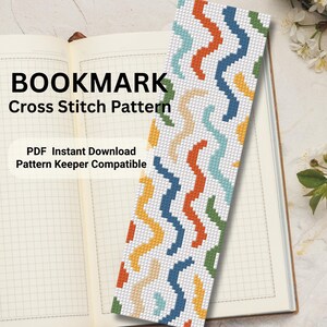 May include: A colorful cross-stitch pattern bookmark with a white background and zig-zag patterns in orange, blue, yellow, and green. The text "BOOKMARK Cross Stitch Pattern" is displayed. The bookmark is on an open notebook.