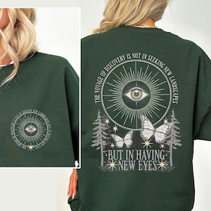 Può includere: Felpa verde scuro con un disegno circolare sul davanti e sul retro. Il disegno include un occhio, raggi di sole, farfalle e il testo "THE VOYAGE OF DISCOVERY IS NOT IN SEEKING NEW LANDSCAPES BUT IN HAVING NEW EYES."