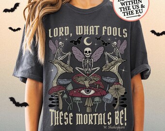 Pode incluir: T-shirt cinzenta escura com uma ilustração de fantasia de esqueletos, cogumelos e borboletas. O texto "LORD, WHAT FOOLS THESE MORTALS BE!" é impresso em estilo gótico, com o nome do autor abaixo. Design artístico único.