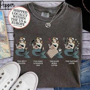Può includere: T-shirt Comfort Colors grigio scuro con quattro illustrazioni di sirene che leggono libri su temi diversi. Il testo sottostante recita: "The Spicy Reader", "The Dark Romance Fan", "The Slow Burn Lover" e "The Fantasy Lover".