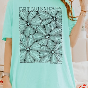 May include: A turquoise blue t-shirt with a black and white graphic of flowers. The text "FARHT LAUGHS IN FLOWERS" is printed on the shirt. The text "Ralph Waldo Emerson" is printed below the graphic.
