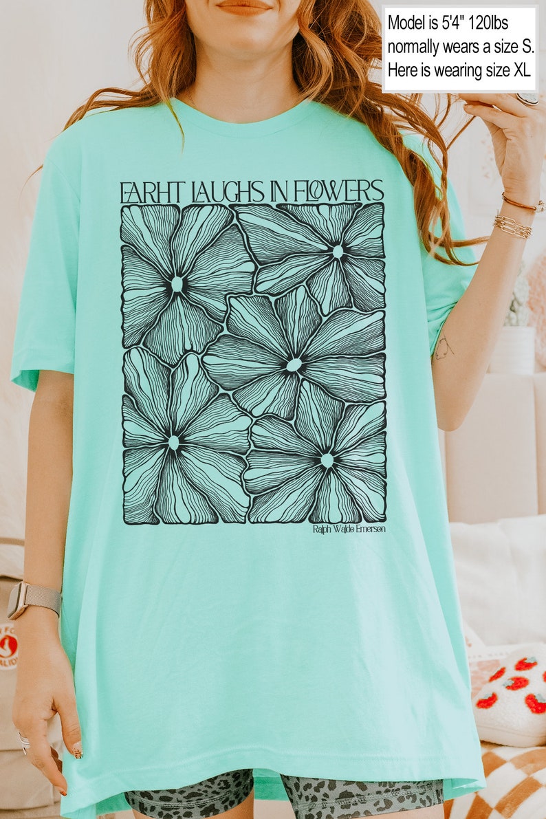 May include: A turquoise blue t-shirt with a black and white graphic of flowers. The text "FARHT LAUGHS IN FLOWERS" is printed on the shirt. The text "Ralph Waldo Emerson" is printed below the graphic.
