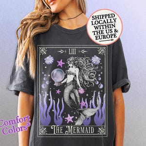Può includere: T-shirt grigio scuro con un'illustrazione dettagliata di una sirena che tiene una sfera di cristallo. Il design include accenti viola, conchiglie e stelle, con le parole "The Mermaid" in basso. La maglietta ha il logo Comfort Colors.