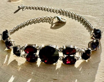 May include: A silver chain necklace with a row of dark red oval-shaped gemstones. The necklace has a lobster claw clasp.