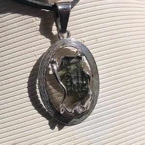 Puede incluir: Un collar con un colgante de plata con una piedra verde, clara y brillante engastada en un marco ovalado de plata. La piedra está engastada en un marco de plata con un diseño ondulado.
