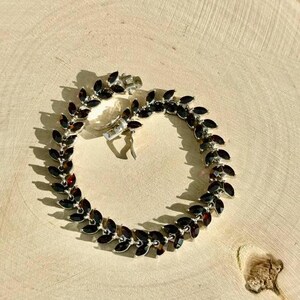 May include: A silver bracelet featuring a row of dark red gemstone accents. The bracelet is made of small, connected links and has a clasp fastening.